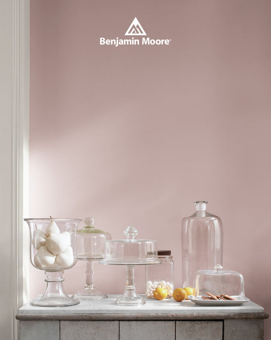 Interior scene featuring a collection of glass cake stands, apothecary jars, and vases on a rustic stone countertop against a soft pink wall. A Benjamin Moore logo is centered at the top.