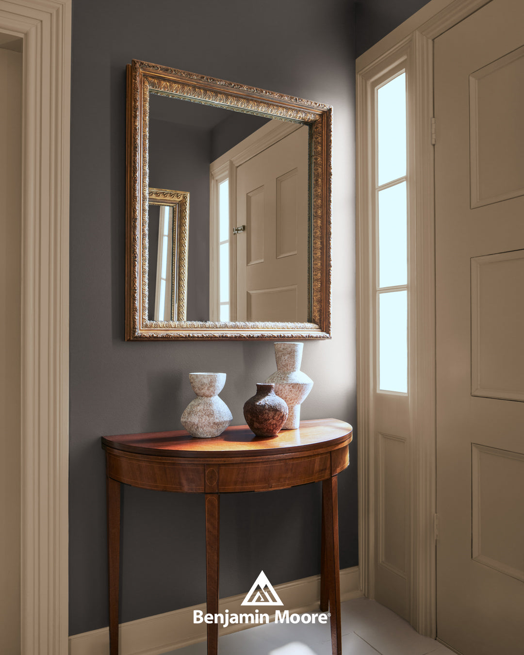 An elegant entryway featuring a dark charcoal accent wall, a wooden half-moon console table with three decorative ceramic vases, and a gold-framed mirror reflecting a white door and hallway. A Benjamin Moore logo is at the bottom.