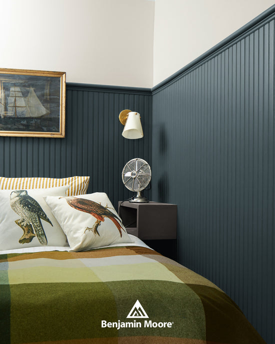 A cozy guest bedroom corner featuring dark teal-blue beadboard wainscoting painted in Benjamin Moore Narragansett Green HC-157. The bed has bird-themed throw pillows and a plaid blanket, with a vintage desk fan and gold wall sconce nearby