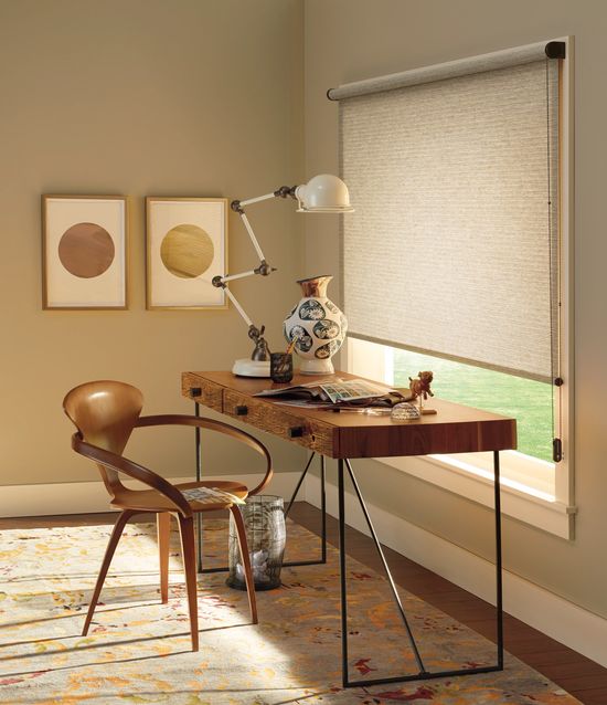 Hunter Douglas Designer Roller Shades in a textured cream fabric installed over a large window in a modern home office with a wooden desk and mid-century chair.