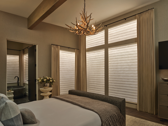 Hunter Douglas Vignette Modern Roman Shades in Belfast Linen fabric, featuring a flat-fold design and paired with side drapery in a luxury primary bedroom with an antler chandelier.