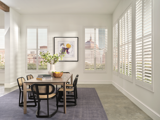 Dining room with wooden table and chairs, framed artwork on the wall, and white Hunter Douglas Palm Beach Polysatin shutters. 