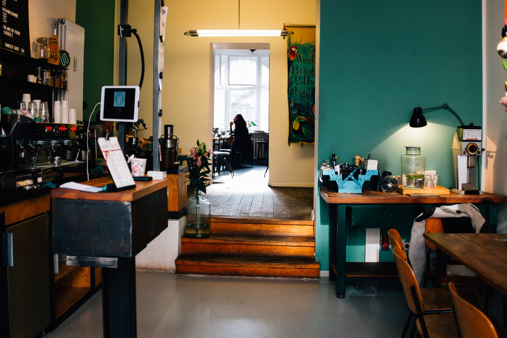 Interior view of a colorful workspace featuring a dark wood coffee bar, a vibrant teal wall with a desk lamp, and a person sitting in a sunlit back room.