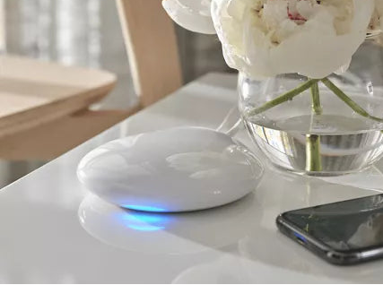 Hunter Douglas Powerview hub on a white lossy table alongside a smartphone and a vase with white flowers. 