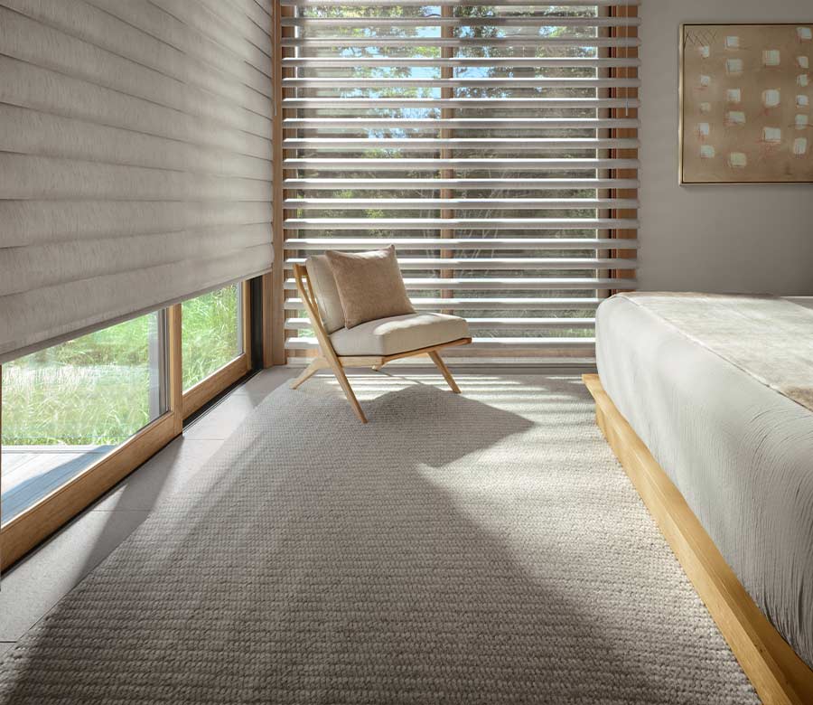 A modern primary bedroom filled with natural light, featuring a low wooden bed frame and a neutral armchair. The large windows are dressed with grey Pirouette Window Shadings, with one shade partially raised to show the horizontal fabric vanes and the outdoor greenery.