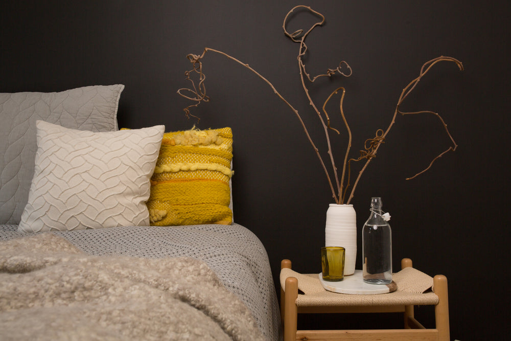 Modern bedroom styling featuring a vibrant yellow textured pillow and grey bedding against a dark charcoal accent wall, paired with a natural wood bedside table and minimalist decor.